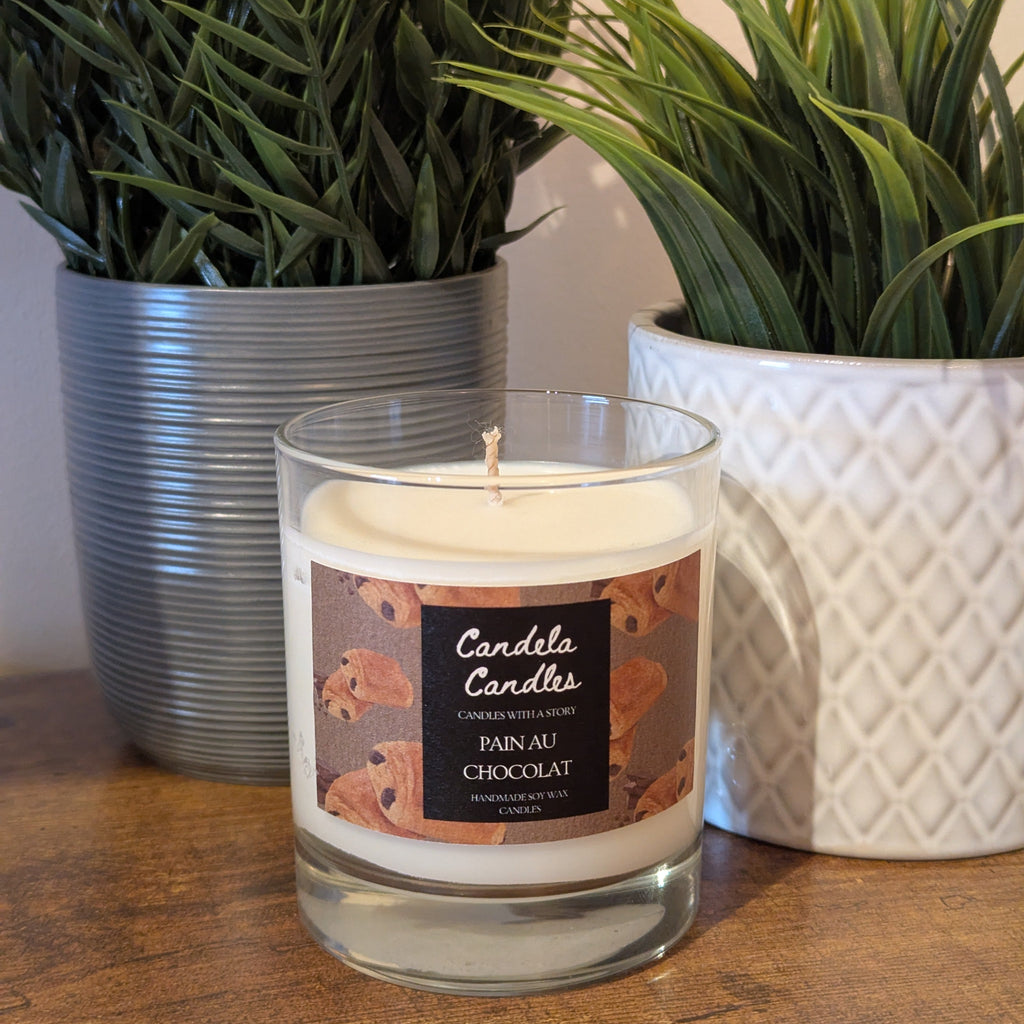 Candle with a label in front of two potted plants on a wooden surface