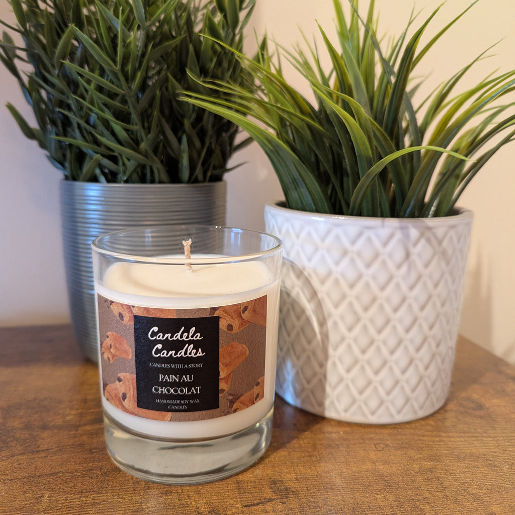 Candle with a label in front of two potted plants on a wooden surface