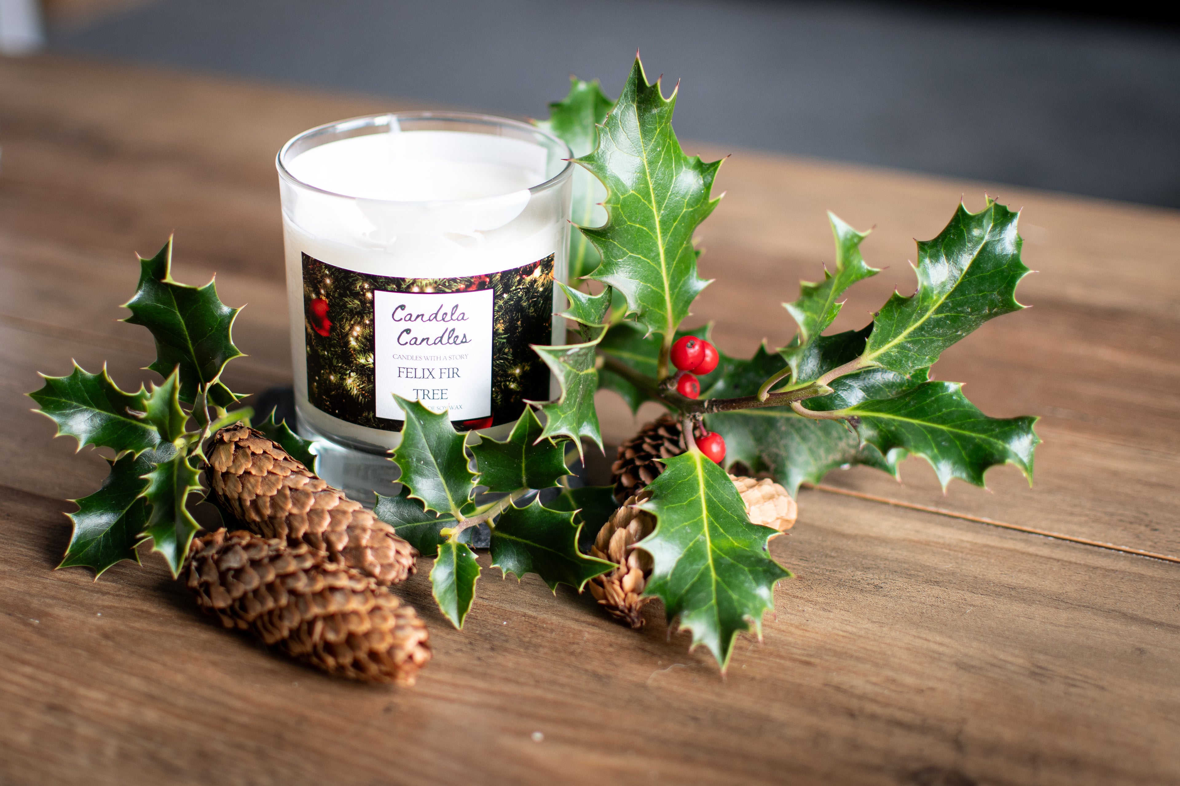 Candle with a label surrounded by holly leaves and pine cones on a wooden surface